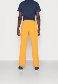 Osoba w musztardowo-żółtych spodniach, granatowym t-shircie i granatowych trampkach Converse stoi tyłem do widza na jednolitym tle.