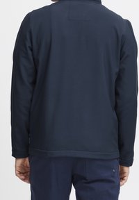 Chaqueta texturizada azul marino con un alto cuello, que presenta un dobladillo recto y puños elásticos. Diseño minimalista; sin logotipos ni patrones visibles.