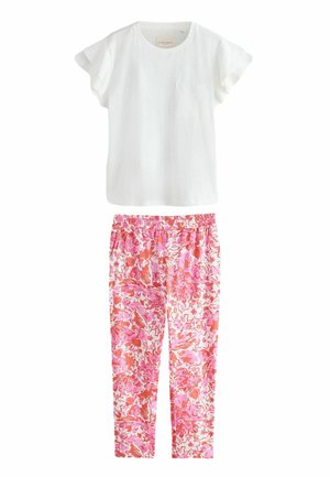 Woman with curly blonde hair wearing cream ruffle-sleeve top and red floral pants standing barefoot in a cozy living room with natural light.