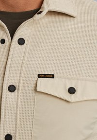 Beżowa koszula w szkocką kratę z teksturowanej tkaniny, z czarnymi guzikami i kieszenią na piersi z napisem "PME Legend" w czerni i pomarańczy.