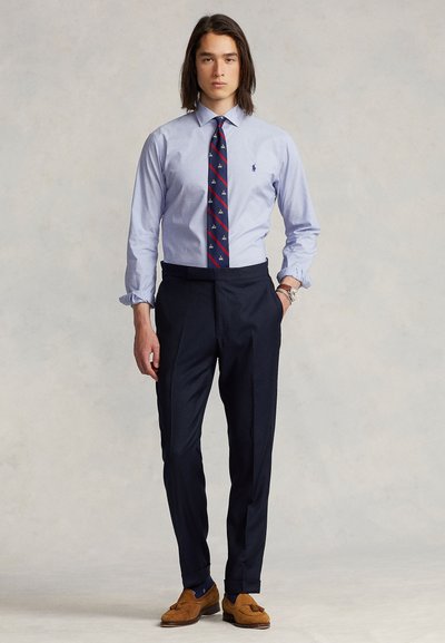 Lys blå skjorte med lange ermer, mørk marinebukser, mønstret slips med rødt og teal, brune loafers, uformell klokke på håndleddet.