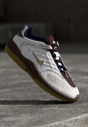 Biały i bordowy but sportowy z gumową podeszwą, logo Nike po boku, przedstawiony unoszący się nad szarą, teksturowaną powierzchnią.