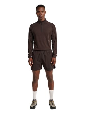 Uomo in piedi che indossa un top marrone scuro a maniche lunghe con zip, pantaloncini abbinati, calzini bianchi a coste e scarpe da trekking beige su sfondo bianco.