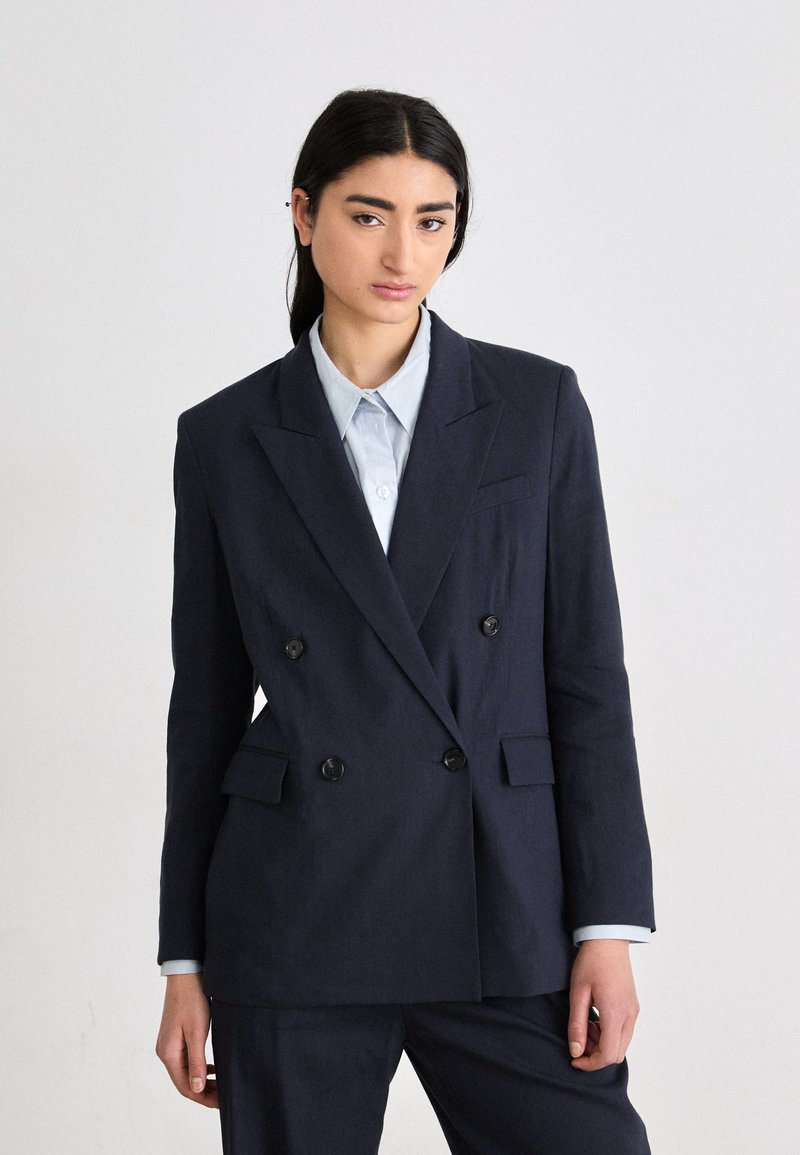 Blazer doppiopetto blu navy in un tessuto testurizzato con rever prominenti, due tasche frontali e bottoni neri, indossato sopra una camicia azzurro chiaro.