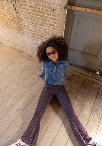 Jonge vrouw met krullend haar en rode zonnebril, zittend op een houten vloer, gekleed in een spijkerjack, met een patroon op haar broek en witte sneakers, tegen een bakstenen muur.