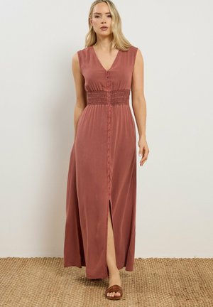 Kobieta o długich blond włosach, ubrana w bezrękawową, kurzowo-różową sukienkę maxi z guzikami z przodu i rozcięciem z przodu, stojąca na plecionej macie.