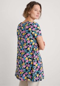 Kobieta z krótkimi włosami, ubrana w kolorową sukienkę w kwiatowy wzór z różowymi, żółtymi i zielonymi liśćmi, stoi na tle jasnego, jednolitego tła.