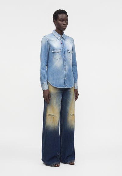 Γυναίκα με ξεθωριασμένο ανοιχτό μπλε τζην πουκάμισο με τσέπες με κουμπιά και φαρδιά τζιν με πορτοκαλί και σκούρο μπλε διαβάθμιση και φθαρμένα γόνατα.