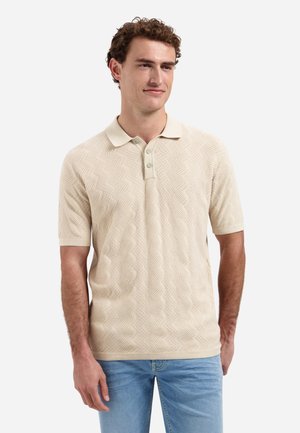 Man met beige, korte mouwen, gestructureerd poloshirt en lichtblauwe spijkerbroek, staande in ontspannen houding tegen een witte achtergrond.