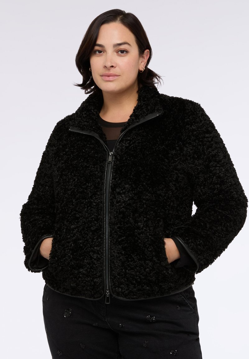 Donna in piedi con le mani nelle tasche, indossa una giacca nera con zip e texture e pantaloni neri con piccoli dettagli in paillettes.