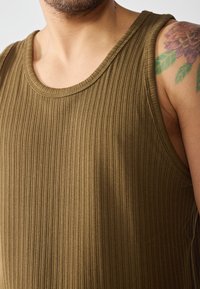 Olíva zöld, bordázott ujjatlan felső széles nyakkivágással, texturált anyagból készült, amely fedi a bőrt és egy színes tetoválást mutat.