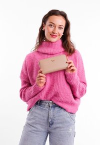 Beige wallet with light polka dots and a gold logo, held in front of a person wearing a pink knitted turtleneck sweater and blue jeans.
