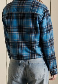 Camicia in flanella a quadratini blu e neri con colletto button-down, dotata di maniche lunghe e taglio dritto, abbinata a jeans in denim blu chiaro.