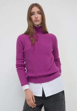 Figurbetonter Ripp-Pullover in leuchtendem Magenta mit hohem Kragen. Kombiniert mit einem weißen Shirt darunter, vor einem neutralen Hintergrund.