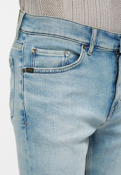 Κοντινό σε ανοιχτό μπλε τζιν με μπροστινή τσέπη με μεταλλικά καρφιά, φερμουάρ και ανοιχτόχρωμο πλεκτό πουλόβερ που είναι μισοβγαλεμένο μέσα.