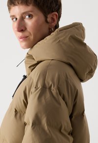 Hnědá péřová bunda s kapucí, která má prošívaný vzor, přední zip a stahovací šňůrku na krku pro nastavitelnou velikost.