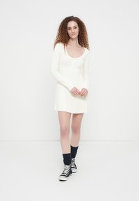 Γυναίκα με σγουρά κόκκινα μαλλιά, που φοράει λευκό πλεκτό φόρεμα, μαύρα ψηλά αθλητικά παπούτσια και σκουρόχρωμες καλτσοδέτες, στέκεται μπροστά σε ένα απλό λευκό φόντο.