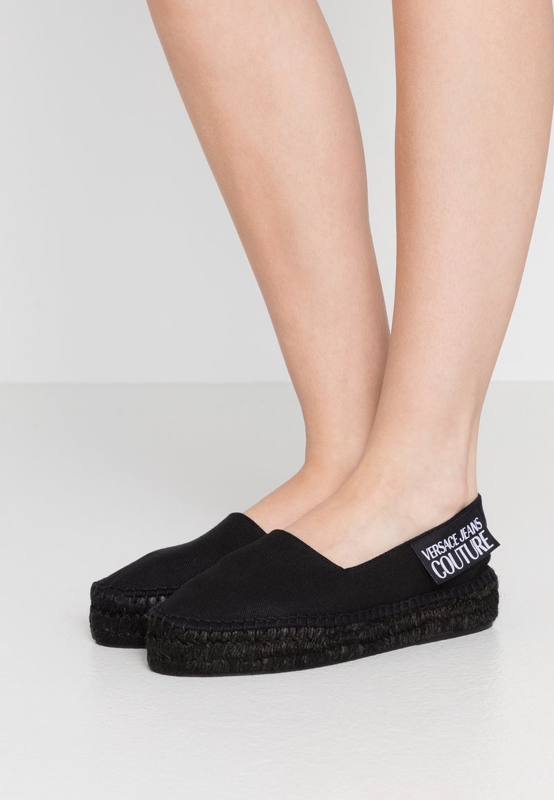 Sorte slip-on espadrillos med flettede platformsåler båret på bare fødder, mærket med "Versace Jeans Couture" label på hælen.