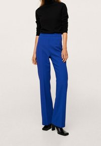 Pantalon bleu sur mesure à jambe droite, en tissu lisse. Associé à un col roulé noir ajusté et des bottines noires à talons.