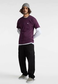 Lila T-Shirt über einem weißen Langarmshirt, kombiniert mit schwarzen Cargo-Hosen und karierten Slip-on-Schuhen. Das Model trägt eine graue Mütze.