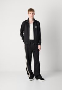Zwarte zip-up jas met witte strepen, met een logo op de borst. Gecombineerd met bijpassende zwarte broek en zwarte sneakers. Gladstof textuur.