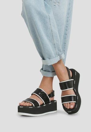 Personne portant un jean bleu clair retroussé et des sandales à plateforme noires avec des bords blancs et un logo "Liu Jo" sur les semelles.