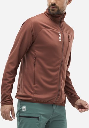Veste zippée rouge avec une finition texturée, dotée de poches latérales et du nom de la marque "Millet" en blanc. Portée avec un pantalon vert clair.