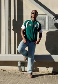 Žalias Adidas polo marškinėlis su baltais rankovėmis ir itališku logotipu, šviesiai mėlynos džinsinės kelnės, rudi batai. Stovi prie betono sienos.