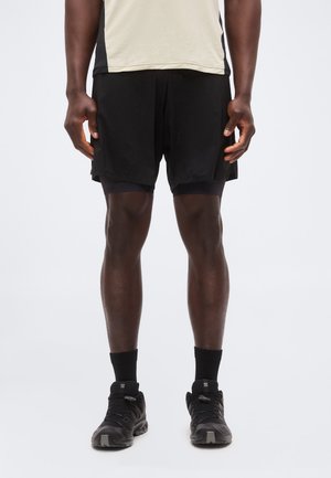 Mężczyzna ubrany w beżowo-czarną sportową koszulkę, czarne warstwowe szorty, czarne skarpetki i czarne buty do biegania, stoi na białym tle.