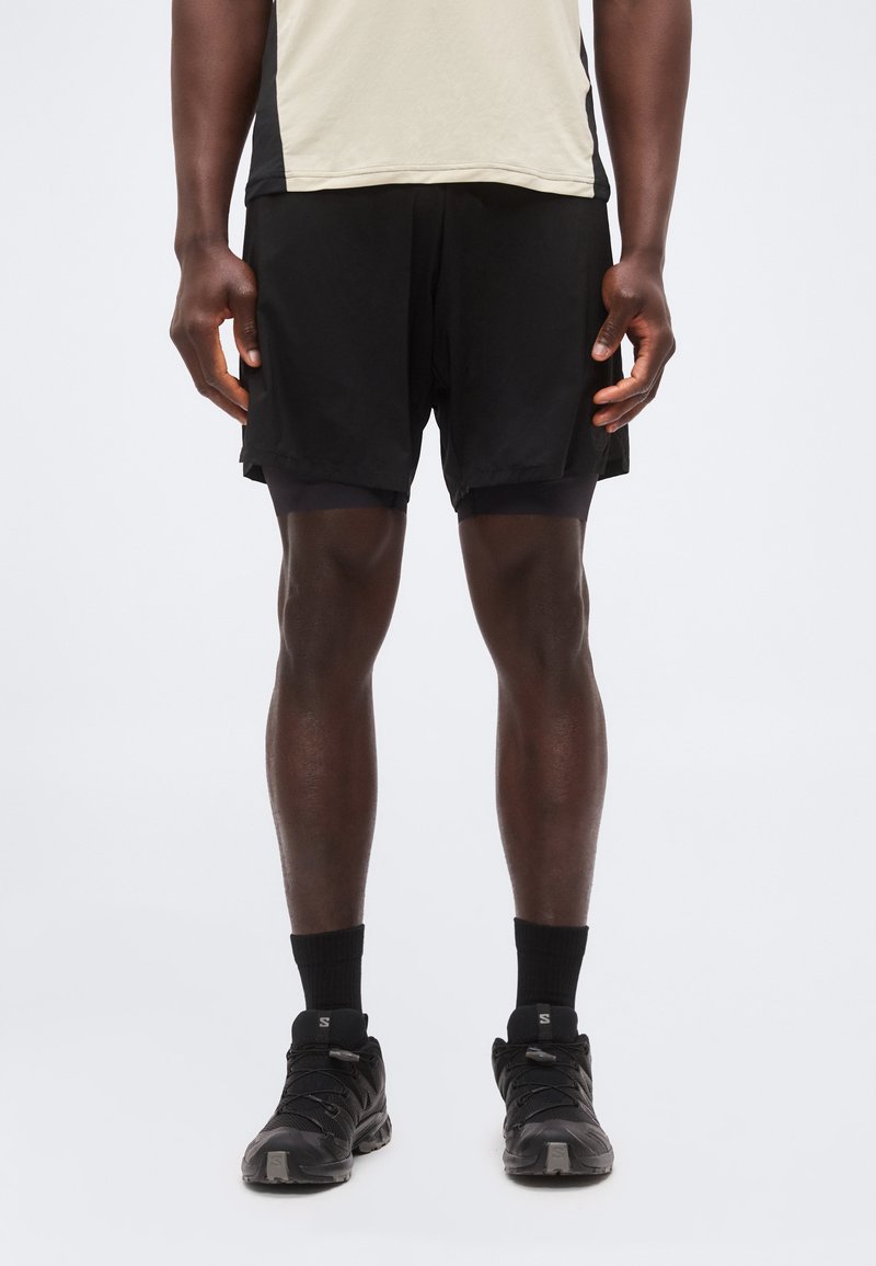 Άνδρας που φοράει μπεζ και μαύρο αθλητικό μπλουζάκι, μαύρο στρωματοποιημένο σορτς, μαύρες κάλτσες και μαύρα παπούτσια για τρέξιμο, στέκεται μπροστά σε λευκό φόντο.