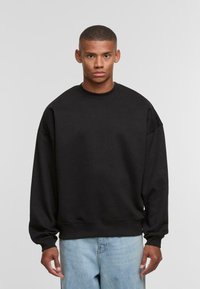 Schwarzes Sweatshirt aus weichem Material, mit Rundhalsausschnitt, überschnittenen Schultern und gerippten Bündchen. Kombiniert mit hellblauen Denim-Jeans.