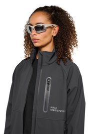 Vrouw met krullend haar die reflecterende sportzonnebrillen en een donkere jas met ritssluiting, borstzak met rits en bedrukte tekst draagt.