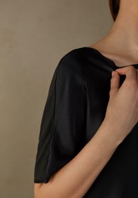 Blusa de satén negro con escote redondo y una textura suave. La manga es holgada y una mano toca suavemente el escote.