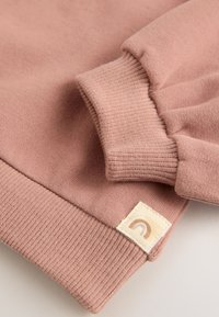 Roze sweater met geribbelde manchetten en zoom, zachte textuur en een klein label met een regenboogontwerp in de onderste hoek.