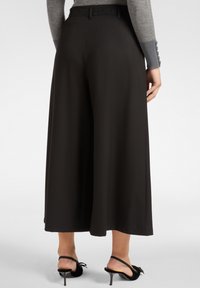 Brede zwarte broek van soepele stof, met een elastische tailleband en een relaxte pasvorm. Gestyled met zwarte hoge hakken.