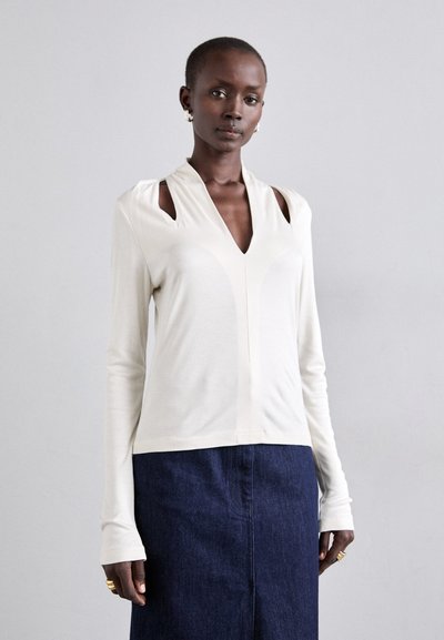 Un haut à manches longues couleur crème avec un décolleté en V profond, des découpes aux épaules et une conception ajustée. Associé à une jupe en denim foncé.