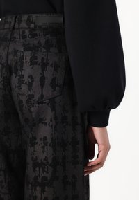 Schwarze Denim-Hosen mit einem dezenten abstrakten Muster, einer Gesäßtasche und einem Etikett am Bund. Das Design ist modern und strukturiert.
