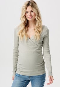 Lichtgroene lange mouwen zwangerschapsblouse met een V-hals en geribbelde textuur, voorzien van zijdelingse plooien voor een aansluitende silhoehette. Gecombineerd met denim jeans.