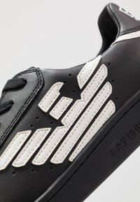 Baskets noires en cuir, avec des rayures diagonales blanches, des détails perforés et des lacets noirs. Logo subtil sur le côté.