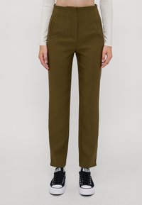 High-waisted, tailored olive green trousers with pleats, side pockets, and ankle-length design, paired with black sneakers.