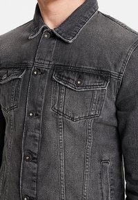 Chaqueta de mezclilla negra con cuello camisero, botones a presión en la parte delantera, dos bolsillos en el pecho y bolsillos laterales. Presenta un acabado desgastado y detalles de costura.