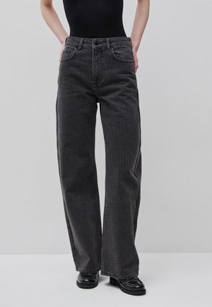 Persona che indossa jeans neri a vita alta e gamba larga con una sottile texture a pois, abbinati a mocassini neri e un top nero aderente.