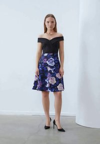 Olkapäiltä avattu mekko, jossa on tiukka musta vartalo ja liehuva, kukkakuosiset hame violetin ja sinisen sävyissä. Mustat korkokengät.