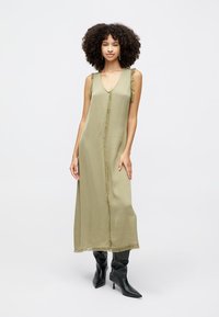 Oliwkowa sukienka bez rękawów z dekoltem w serek, z postrzępionymi brzegami i prostym krojem. Modelka nosi czarne kozaki za kolano.