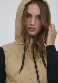 Beige gesteppte Weste mit Kapuze, ausgestattet mit einem Reißverschluss und schwarzen Akzenten an den Seiten. Weicher, leichter Stoff mit glatter Textur.