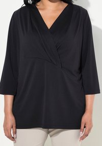 Musta, kolmanneksen mittainen hiha-toppi, jossa on V-pääntie; sisältää roikkavaa kangasta ja kevyen kiertoyksityiskohdan edessä. Pehmeä, sileä rakenne.