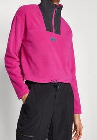 Polaire à enfiler de couleur rose vif avec un col gris, fermeture à moitié zippée et accents contrastants bleu sarcelle. Design court avec ourlet à cordon de serrage.