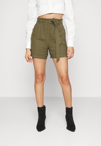 Pantaloni corti verde oliva con cintura elastica e dettaglio di fiocco frontale, realizzati in un materiale testurizzato, abbinati a stivaletti neri alla caviglia.