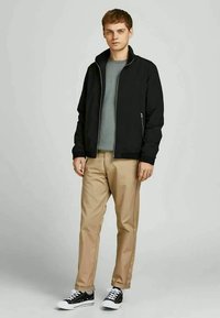 Black lightweight jacket with a high collar, gray t-shirt underneath, khaki pants, and black sneakers with white soles.