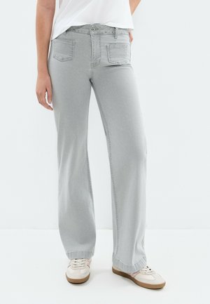 Persona vistiendo vaqueros de pierna ancha de color gris claro y zapatillas blancas con detalles en rosa, de pie frente a un fondo blanco liso.